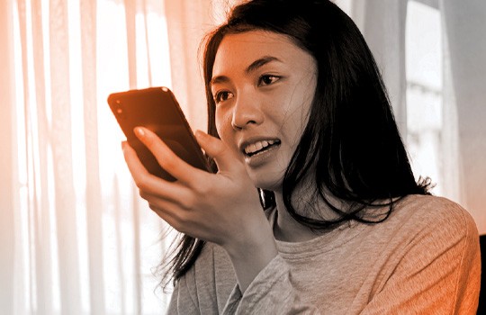 This photo shows a woman holding her mobile phone in her hand. The phone is held in front of her face, near her mouth. She is talking at the phone as if to ask the phone’s integrated virtual assistant a question. The photo suggests that online merchants must optimize for multilingual voice search if they want their products to be found on the crowded digital shelf. 