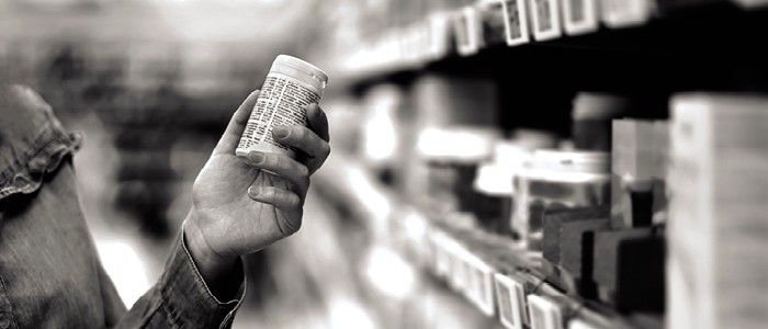 A hand holds a bottle of pills in a pharmacy aisle.