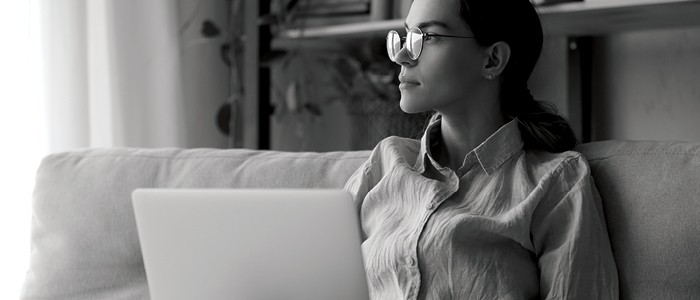 Woman wearing glasses and sitting on couch looks away from her laptop